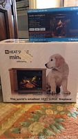 Photo of the boxed Heat Surge Mini Glo electric fireplace featuring the puppy image and claim as world's smallest Heat Surge fireplace with logo and product photo.