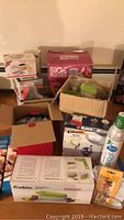 Photo showing assortment of boxed kitchen appliances including Magic Bullet, Black & Decker mixer, baking mats, and various boxed and bagged kitchen tools in cardboard boxes.