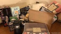 Overview photo showing boxed steam brush, laundry items, bulbs, and blankets in cardboard box and surrounding area.
