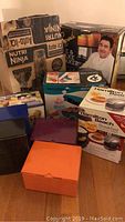 Boxed Nutri Ninja Auto-IQ blender, Emeril pie and cake maker, Hamilton Beach breakfast maker, Yes Chef waffle makers and other small appliances in original boxes clustered together on floor.