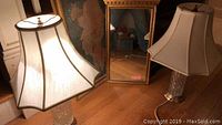 Photo showing two cut glass lamps with beige and gold trim shades, gold framed mirror standing behind, and framed wall art leaning against wall.