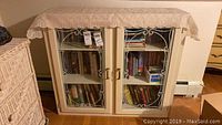 Front view of white bookcase covered with lace cloth showing mostly front and top edges, glass doors reveal books stored inside.