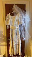 Full view of vintage wedding dress hung on door with lace sleeves and tiers, veil draped on side.