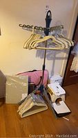 Drying rack with three arms holding multiple plastic hangers, pink fabric laundry hamper bag visible behind, boxes and other small items on floor