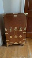 Front view of upright decorative cabinet showing gold-toned metal hardware on corners, handles, and hinge plates