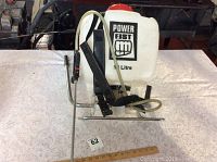 Front view showing white plastic tank with black Power Fist logo and 15 litre capacity, red cap on top. Straps and metal frame visible along with spray wand and hose.