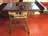 Front view of Rockwell/Beaver 10 inch table saw showing whole metal table surface, saw blade slot missing center piece, metal stand, and general wear.
