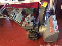 Side view of snowblower showing engine, handlebar, chute and auger housing on red floor