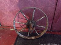 Old steel wagon-type wheel seen from front, showing spokes and axle mount, rusty and heavily aged metallic surface.