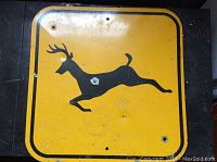 Front view of the square yellow metal road sign with a black silhouette of a running deer showing overall condition and mounting holes.