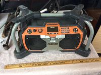 Front view of Ridgid contractor radio with keypad, knobs, dual speakers, and metal protective frame on a table with measuring ruler in front.
