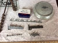 Lot contents including Pontiac emblem, two Laurentian emblems, Safari emblem, Chevrolet hubcap, and Vintage First Aid Travel Kit on lace fabric.