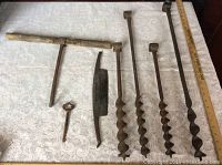 Full view of all seven items on lace tablecloth including four hand forged iron drill bits, forged blade, hook with wooden handle, and old key