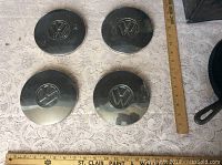 Four Volkswagen hub caps laid out flat with wooden rulers showing size approximately 5.5 inches diameter.