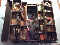 Top view of open vintage metal tackle box showing wooden fishing lures, fishing hooks, and various fishing accessories arranged in compartments separated by metal dividers and cork liners.