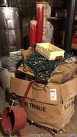 Wide view of large bundle of Christmas string lights piled on a cardboard box with pair of tall red decorative candles in background