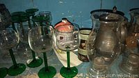 Clear glasses with green stems arranged on a shelf along with ceramic and pewter teapots in the background