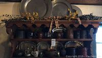 Wooden shelf display with two large pewter plates on top shelf, middle shelf holds three pewter platters and stoneware crocks, bottom shelf shows wooden dough bowl, ceramic crock and small decorative pieces