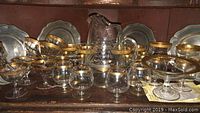 Glassware set with various stemmed glasses and bowls featuring gold patterned rims, arranged on a wooden surface.