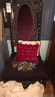 Photo of the carved wooden chair showing the oval burgundy upholstered back and floral upholstered seat with a red decorative pillow placed on it.