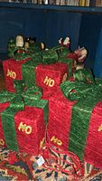 Six red glitter light-up gift boxes with green bows and yellow 'HO' lettering arranged in a cluster with two Santa figures and nutcracker dolls in background