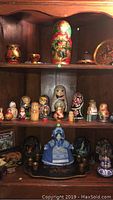 Wooden cabinet shelf displaying nesting dolls, lacquer cups, covered bowl and dish; central large red floral matryoshka