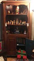Wood corner cabinet featuring three open shelves on top holding multiple nesting dolls and figurines, and a closed two-door cabinet underneath with two shelves inside.