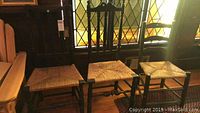 Three wooden chairs shown together in front of stained glass window with visible dark finish wood frames and woven rush seats.