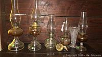 Full view of five assorted oil lamps and clear glass vase on dark wood shelf showing varying sizes and styles.