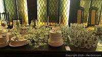 Wide view of large collection of clear glasses with leaf designs arranged on table along with white dinnerware plates and bowls stacked nearby.