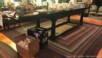 Full view of the long heavy wooden dining table with multiple ornate legs over a patterned rug, some boxes placed nearby.