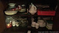 Dark cabinet shelf filled with various bone china plates, cups, saucers, glassware, and a teapot, showing the collection's overall contents and condition.