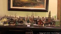 Full display of the Christmas village with multiple lighted buildings set up on a table showing various styles and sizes of miniature houses along with trees and a large Christmas tree in center.