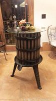 Full view of the wine press table with round glass top, metal corkscrew, and plastic grapes on top, showing wooden slat base and three cast iron legs.