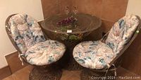 Photo showing round glass top table on wicker base with two matching padded floral chairs, two wine glasses, and plastic grapes on the table