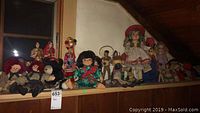 Wide shelf view containing about 15 dolls and three stuffed animals displayed in a row under a slanted ceiling.