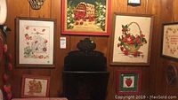 Wall with multiple framed strawberry themed needlepoint artworks, various sizes from 5x6 to 12x18 inches, arranged above and beside a dark stove.