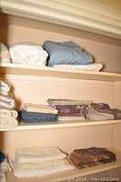 Shelf with folded linens including white, beige, blue, and patterned sheets and pillowcases.