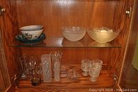 View of upper wood cabinet shelf showing three bowls: blue ceramic salad bowl with fruit pattern, frosted glass bowl with flower pattern, yellow scalloped glass decorative bowl. Lower shelf holds various wine glasses, two vases, small pottery pieces, and glass leaf bowl.
