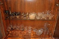 Photo showing an assortment of clear cut glassware including a pitcher, two tumblers, various wine glasses, small bowls, and dishes on wooden shelves.