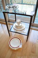 View of Tuscan Louise china set displayed on metal table and stacked plates on floor, showing teacups, plates, and silverware on top shelf