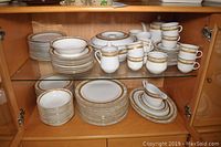 Two-tier cabinet with upper shelf showing bowls, plates, cups, coffee pot and cream & sugar; lower shelf showing stacks of bowls, plates and a gravy boat on plates
