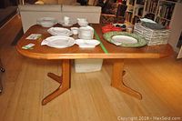 Front side view of the wooden dining table showing the two T-shaped legs and the rounded rectangular tabletop with dishware placed on top for scale and context.