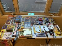 Photo showing an assortment of packaged tools including wrench, clamps, sanding discs, protractor, screws, bits and wire strippers arrayed on a table near a window.