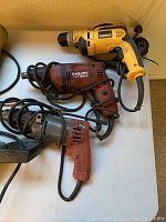 Three corded electric drills from DeWalt, Hilti, Milwaukee brands on a white table surface, showing body and cords.