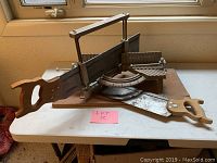 Full view of vintage metal miter box with attached handsaw and wooden handle on folding table.