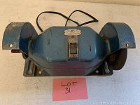 Front view showing the blue metal body, grinding wheels, safety guards, and the on/off switch of the bench grinder.