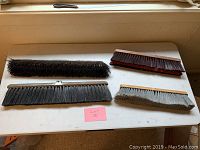 Four large brushes on a white table, showing various bristle materials and colors, all with wooden bases. Brushes are arranged with a pink lot label visible.