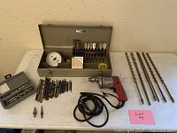Overview of all items including Milwaukee drill, metal case with drill bits, loose drill bits and auger bits arranged on table