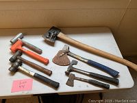 View of lot with seven axes and hammers including large axe with sheath on blade, hammer with orange handle, and smaller hammers and hatchets on white table.
